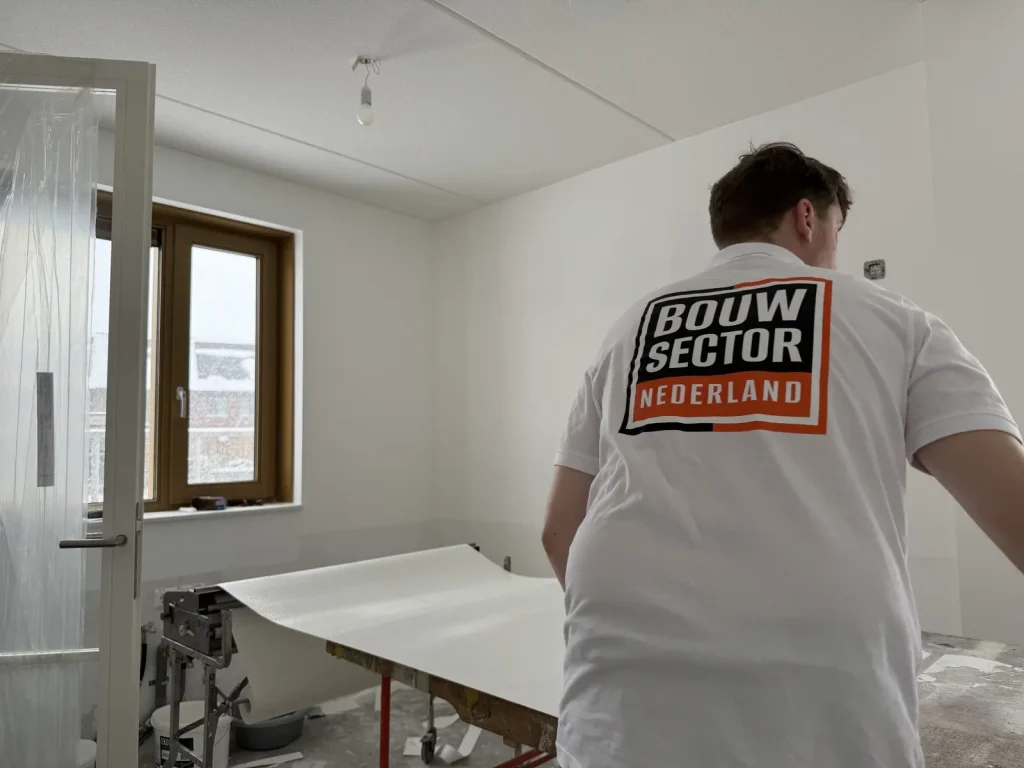 Vakman in wit werkshirt legt een baan vlies klaar op de plaktafel naast het raam