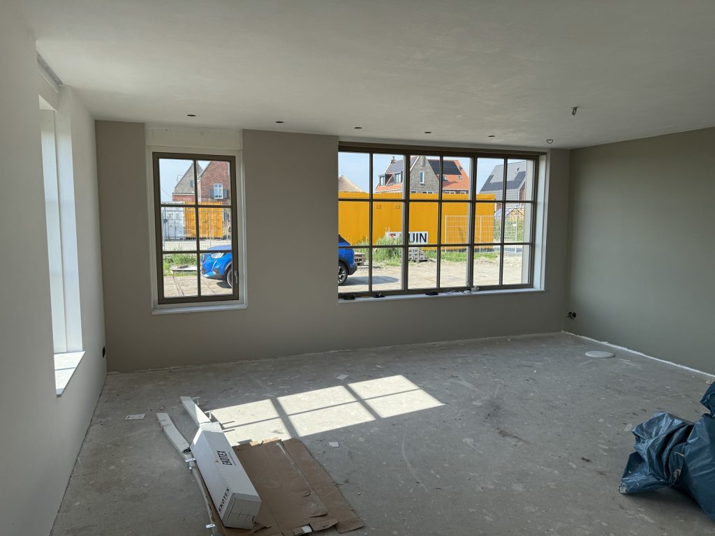 Nieuwe woonkamer met grote roedenramen en egaal afgewerkte wanden in een warme grijstint